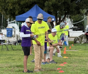 Kids participating in a fishing clinic outdoors, as experienced guides in bright shirts teach them how to cast rods on a grassy field with cones set up for practice.